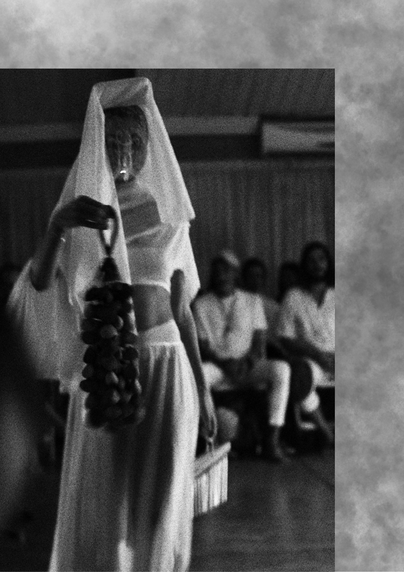 Veiled figure in white holding a beaded strand before a seated audience in grainy black and white.