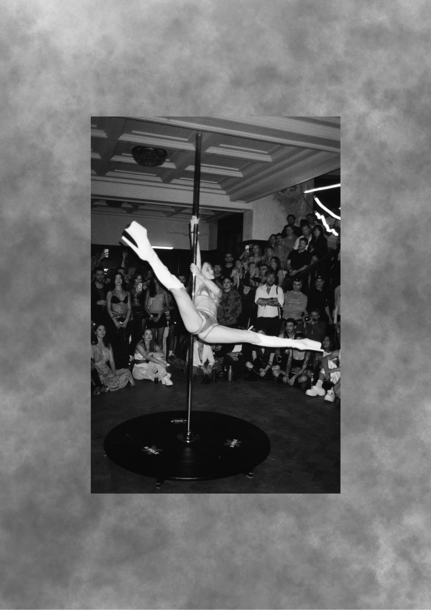 Aerial pole performer in a horizontal split before a seated audience.